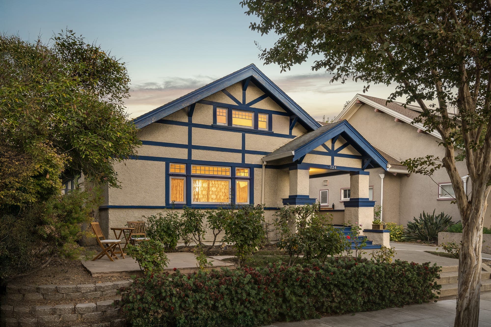 The Craftsman bungalow at dusk, its gabled porch and windows lit warm against a soft evening sky.
